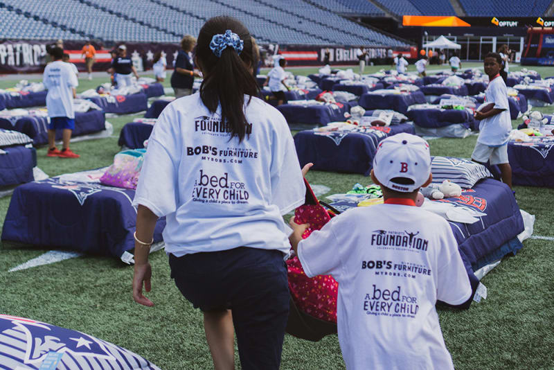 A woman and child wearing white t-shirts with "FOUNDATION," "BOB'S FURNITURE," and "a bed FOR EVERY CHILD" text, walking hand-in-hand on a football field with multiple beds arranged, promoting a charity event for providing beds to children.