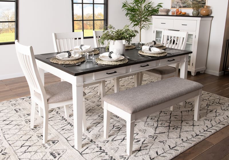 Dining room set with a white wooden table featuring a dark wood top, four white cushioned chairs, and a beige upholstered bench on a patterned rug.