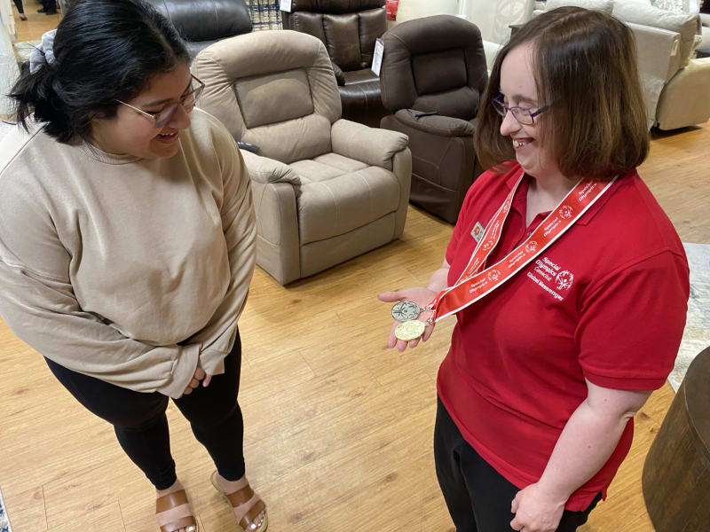 Two women standing indoors on a light wood floor near beige and dark brown recliner chairs. One woman wears a red polo shirt with "Special Olympics Georgia Global Messenger" text and holds two medals with red ribbons around her neck. The other woman wears a beige sweatshirt, black pants, and brown sandals.
