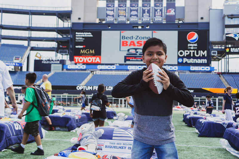 A child holding a football on a stadium field with Patriots-themed pillows and stuffed animals. Stadium scoreboard displays "Bed-Zone BASH" and sponsors Toyota, Pepsi, Optum, and Putnam.
