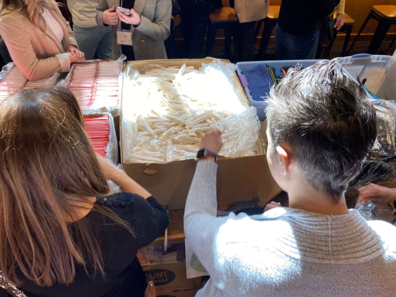 Two people sorting and packing individually wrapped white and red plastic straws into large cardboard boxes, likely for distribution or event preparation.