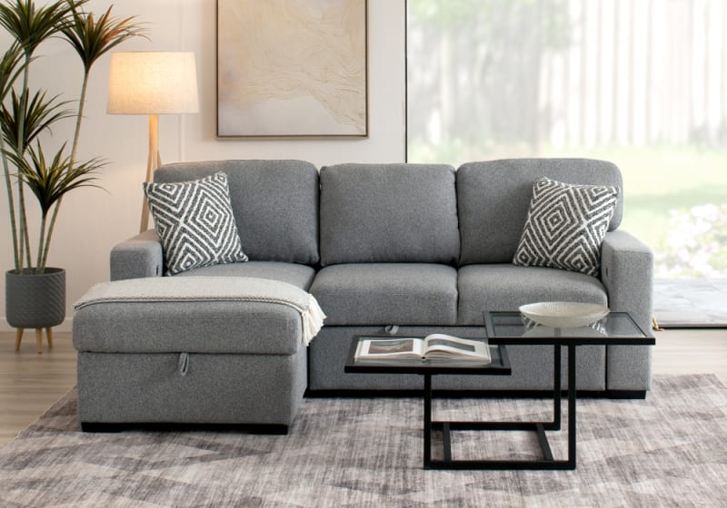 Gray fabric sectional sofa with chaise, geometric patterned throw pillows, glass nesting coffee tables, floor lamp, and indoor plant in modern living room.