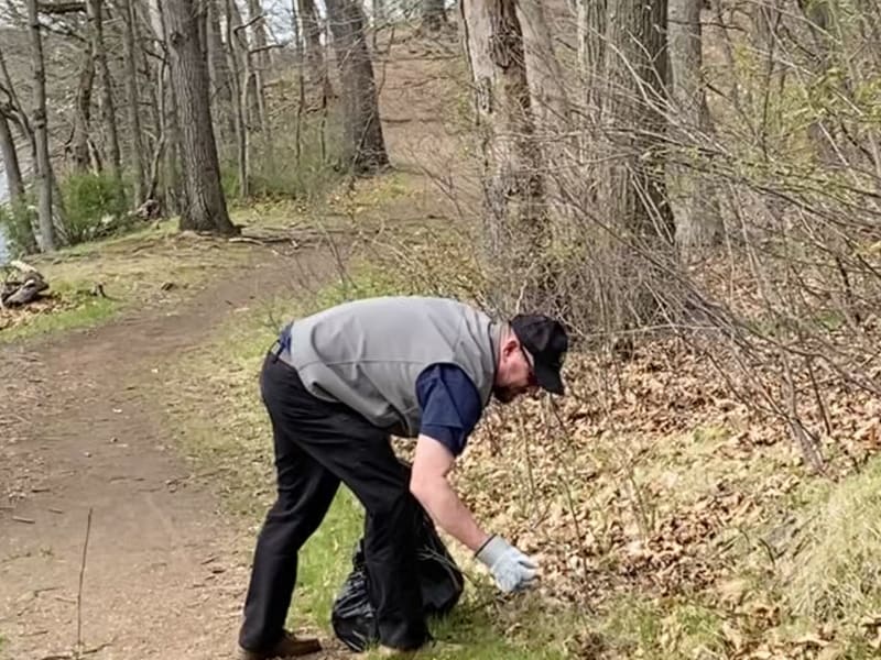 A man wearing a gray vest, black pants, and gloves is picking up trash in a wooded park area along a dirt trail, promoting environmental cleanup.