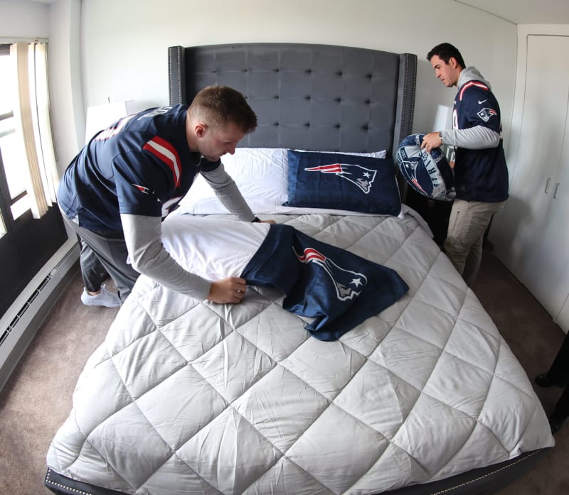 Two people in New England Patriots jerseys arranging matching Patriots-themed bedding, including a pillow and blanket, on a gray upholstered bed in a bright bedroom.