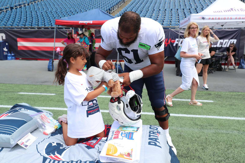 A football player in a white Patriots jersey with number 61 interacts with a young girl kneeling on a Patriots-themed blanket on a stadium field. The player holds a white football helmet while the girl holds a stuffed animal. The background shows empty blue stadium seats and tents with Patriots branding.