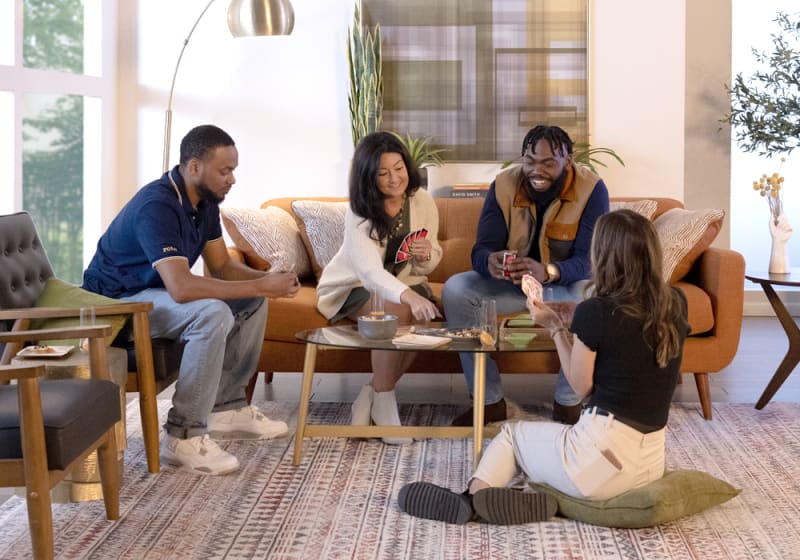 Four people playing cards in a modern living room with an orange sofa, glass coffee table, patterned rug, and natural light.