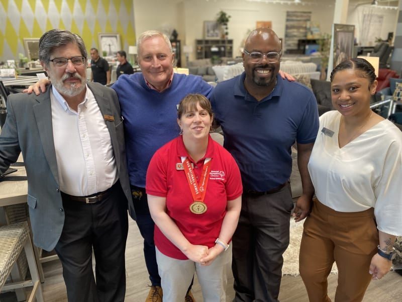 Five people standing indoors in a casual group pose, with one person wearing a red polo shirt and a gold medal on a red lanyard, surrounded by others in business casual attire.