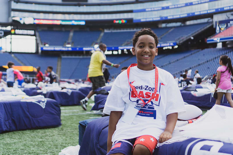 A child wearing a white t-shirt with red and blue text "Red-Zone BASH" and a red lanyard, sitting on a bed in a large stadium filled with similar beds and other children.