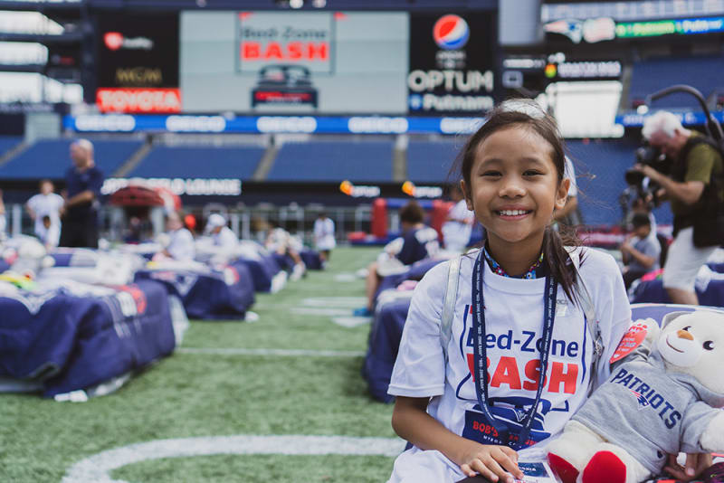 A young girl wearing a white "Red-Zone BASH" t-shirt and lanyard sits on a football field holding a Patriots teddy bear, with rows of bags and people in the background at a stadium event.