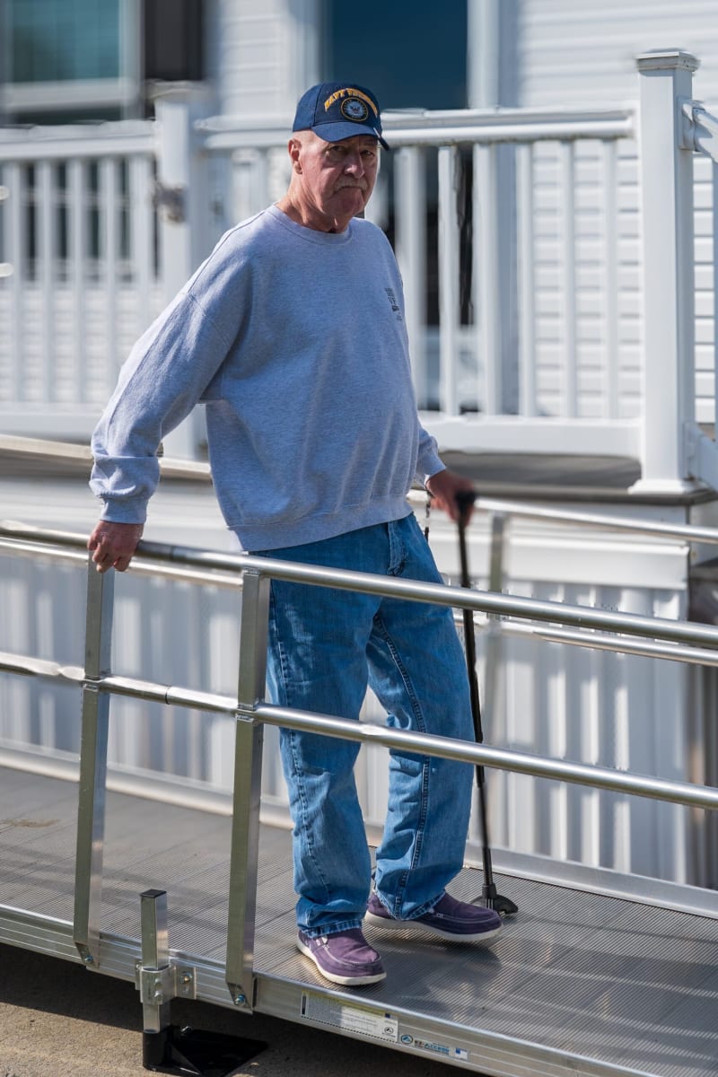 Man wearing a navy blue Navy baseball cap, gray sweatshirt, blue jeans, and purple sneakers, using a black walking cane on a metal ramp with railing.