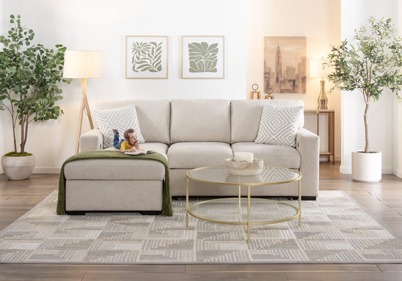 Beige fabric sectional sofa with matching ottoman and patterned cushions in a modern living room setting. Includes glass-top gold metal coffee table, decorative plants, and neutral area rug.