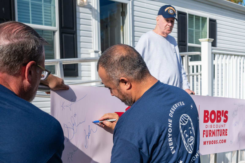 Three men signing a large white banner with blue ink signatures in front of a house. The banner features red text: "BOB'S DISCOUNT FURNITURE MYBOBS.COM." One man wears a navy blue shirt with a logo and text "EVERYONE DESERVES" around a smiling face graphic.