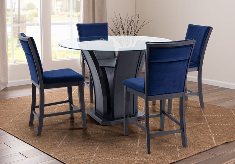 Modern round glass dining table with a black wooden pedestal base, paired with four navy blue velvet upholstered chairs on a brown woven rug.