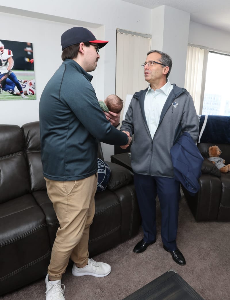 Two men shaking hands indoors, one holding a baby wrapped in a green blanket. Both wear casual jackets; one in navy and khaki pants, the other in a gray hoodie with a Patriots logo. The setting includes leather sofas, a window with vertical blinds, and a teddy bear on a chair.