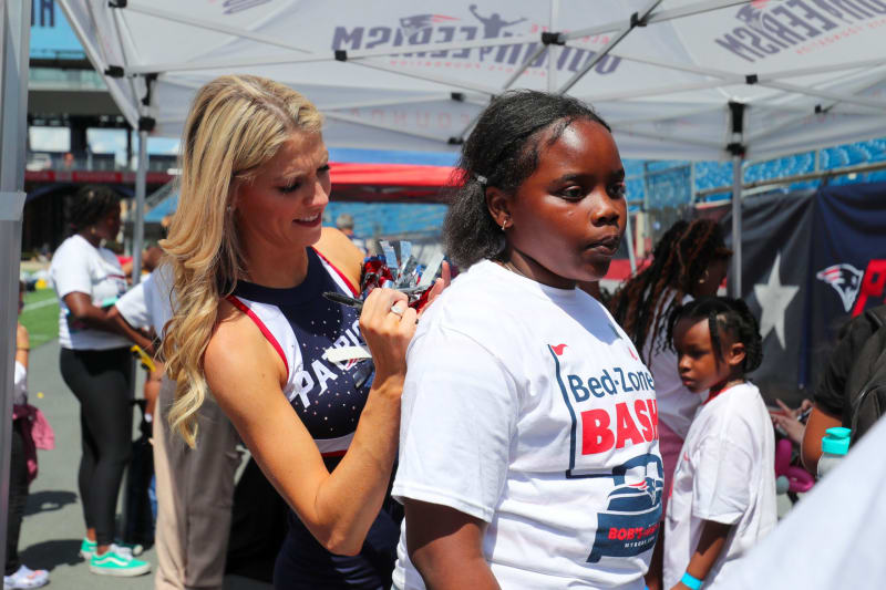 A blonde cheerleader in a navy and white Patriots uniform signs an autograph on a fan’s white "Bed-Zone Bash" t-shirt under a tent at a sports event.