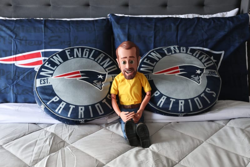 Doll wearing a yellow shirt and blue jeans seated on a bed with New England Patriots pillows and bedding featuring the team logo and colors.