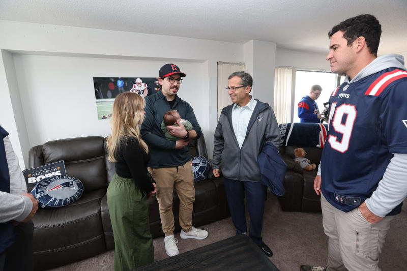 Group of people gathered in a living room with New England Patriots decor, including pillows, a blanket, and a teddy bear wearing a Patriots shirt. One person holds a baby, and another wears a Patriots jersey with number 19. A football-themed poster is on the wall.