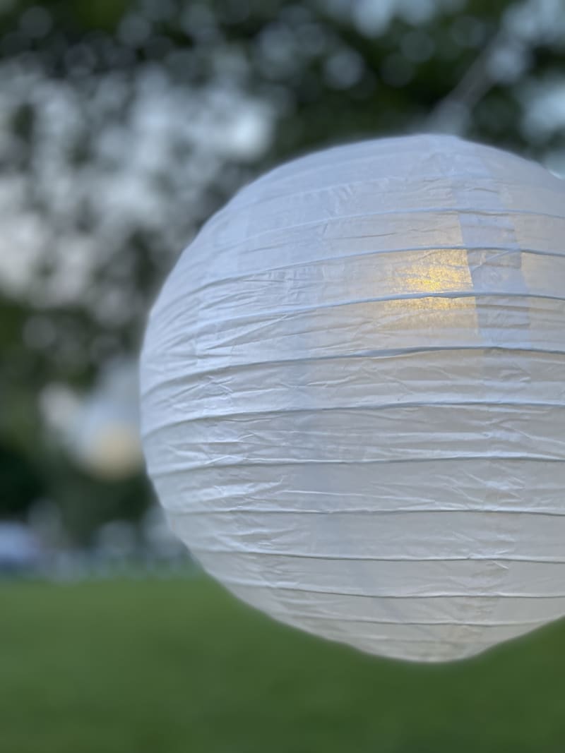 White paper lantern with a warm light inside, hanging outdoors with blurred green trees and grass in the background.