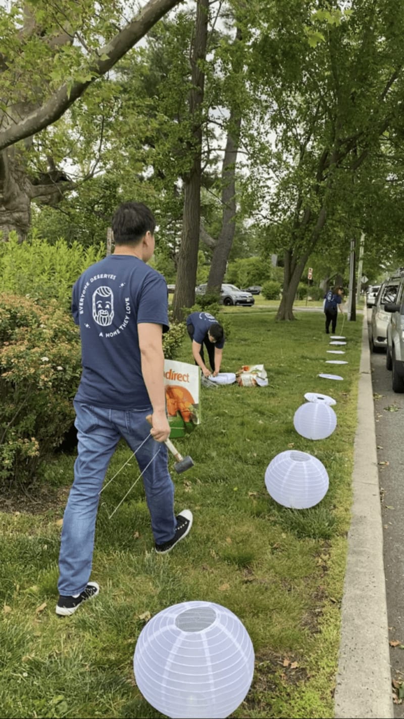 A person holding a mallet and stakes sets up white paper lanterns along a grassy sidewalk, with two others arranging items nearby.