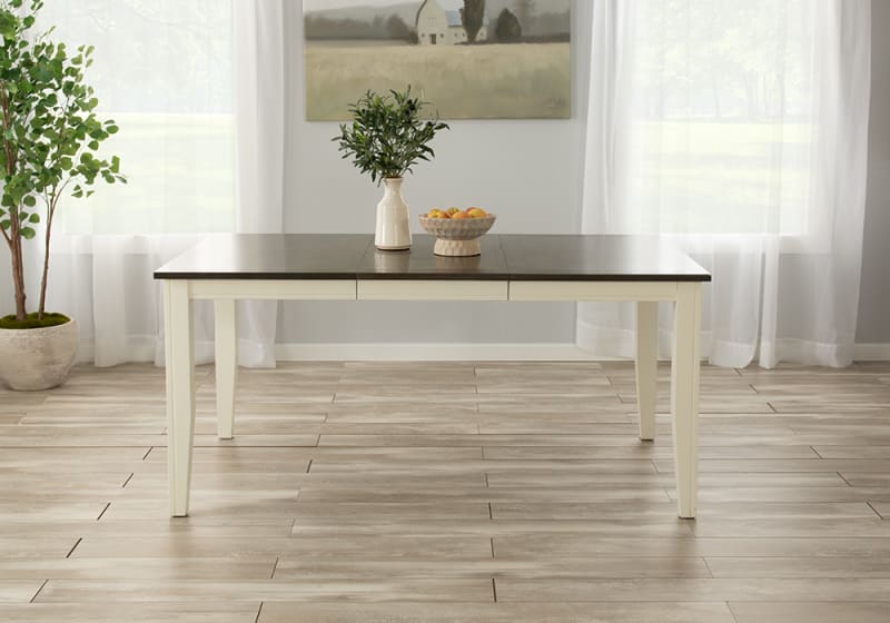 Rectangular dining table with dark wood top and cream-colored legs, styled with a ceramic vase holding green foliage and a beige bowl of fruit, set in a bright room with light wood flooring and sheer white curtains.