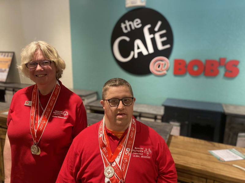 Two individuals wearing red Special Olympics Connecticut Global Messenger shirts and medals stand inside a café with a sign reading "the Café @ BOB'S" on a teal wall behind them.