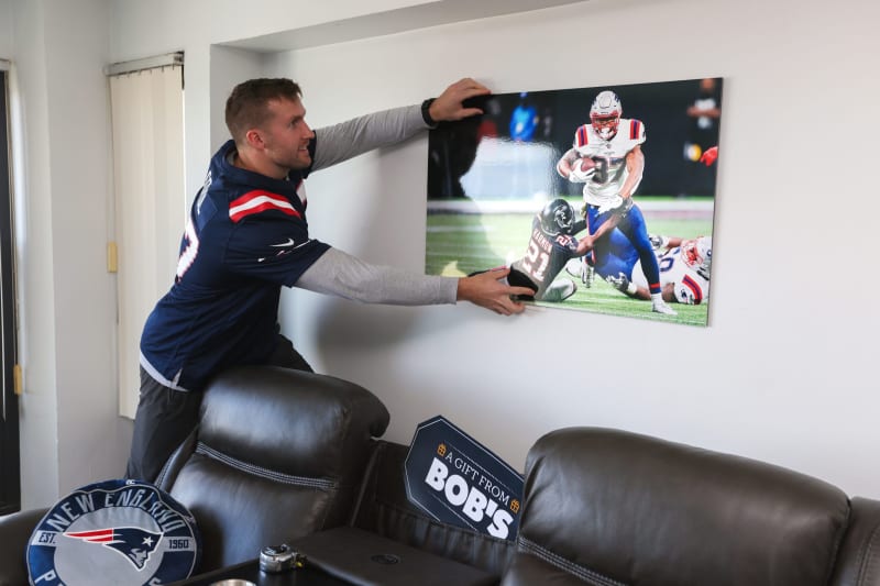 Man wearing a New England Patriots jersey hanging a framed football action print on a white wall, with a Patriots pillow on a leather couch nearby.