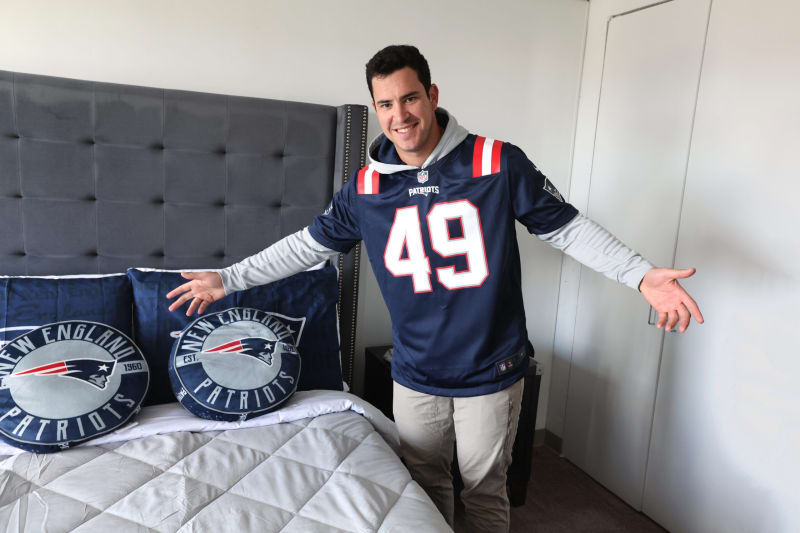 Person wearing a navy blue New England Patriots NFL jersey with number 49, standing next to a bed with matching Patriots pillows and gray bedding.