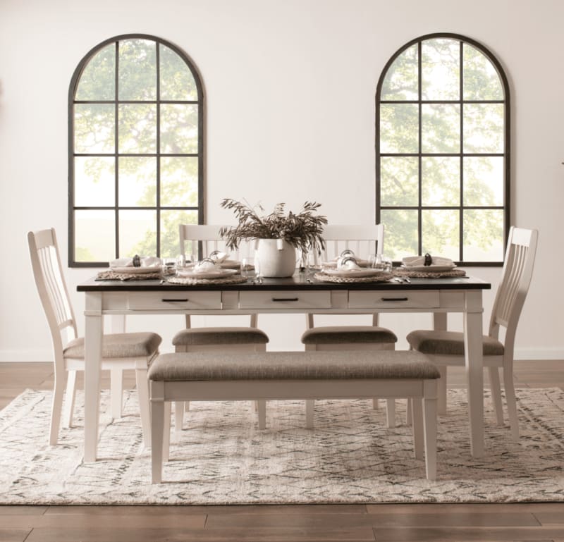 Dining room set with a black tabletop, white wooden frame, beige cushioned chairs, and a matching bench on a patterned rug, under arched windows.
