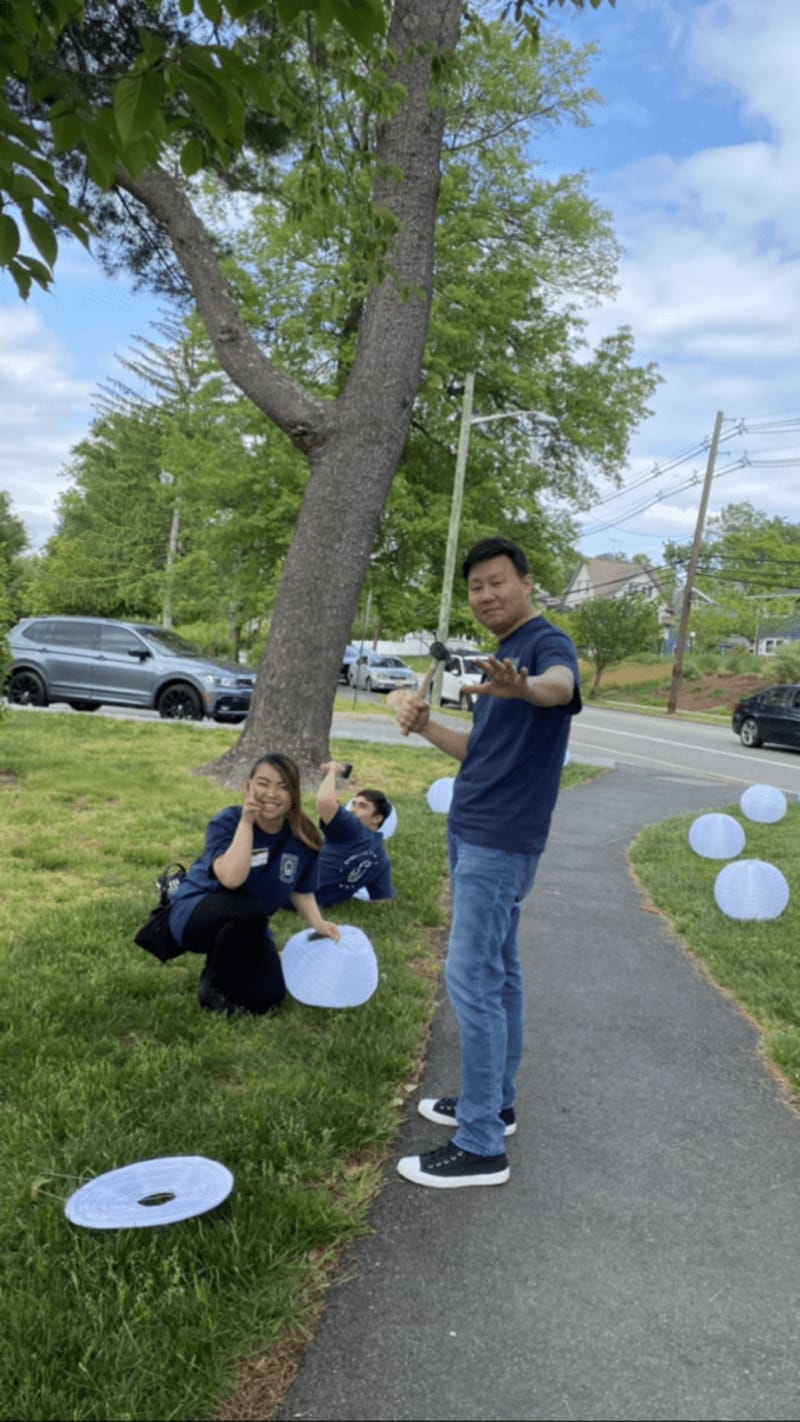 Three people outdoors on a grassy area near a sidewalk, holding white paper lanterns, with trees, parked cars, and a street in the background.