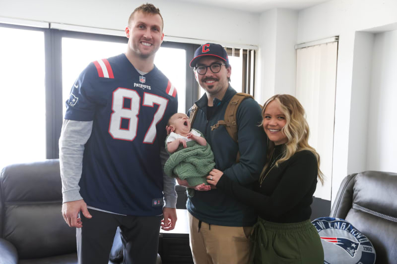 A man wearing a navy blue New England Patriots jersey with number 87 stands next to a couple holding a baby wrapped in a green blanket inside a bright room with leather chairs.
