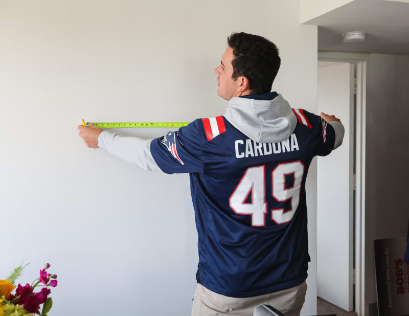 Person wearing a navy blue New England Patriots jersey with "CARDONA" and number 49, measuring a wall with a tape measure.
