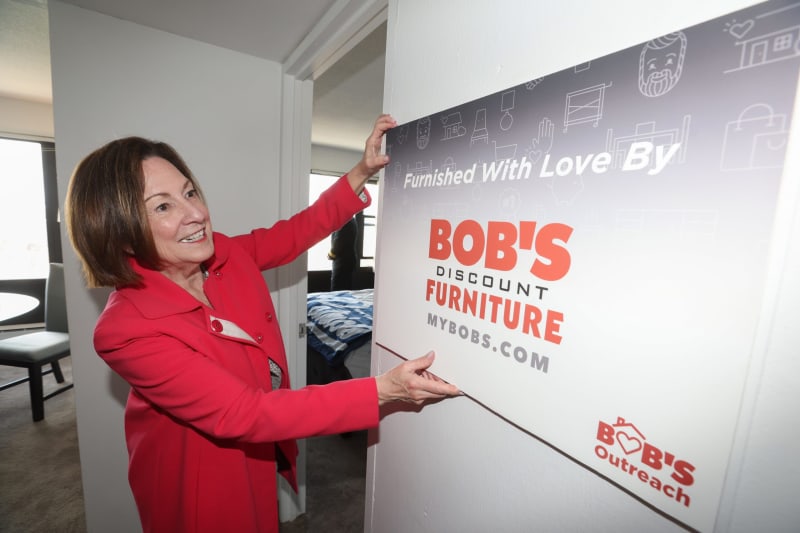 Person in red jacket holding a sign that reads "Furnished With Love By BOB'S Discount Furniture MYBOBS.COM" with BOB'S Outreach logo, promoting furniture brand and outreach program.