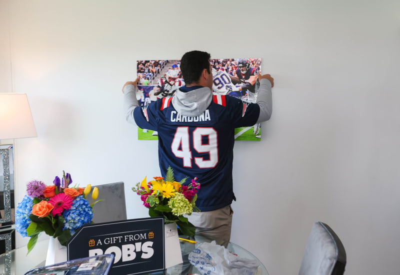 Man wearing a navy blue New England Patriots jersey with "Cardona 49" hangs a framed football action photo on a white wall.