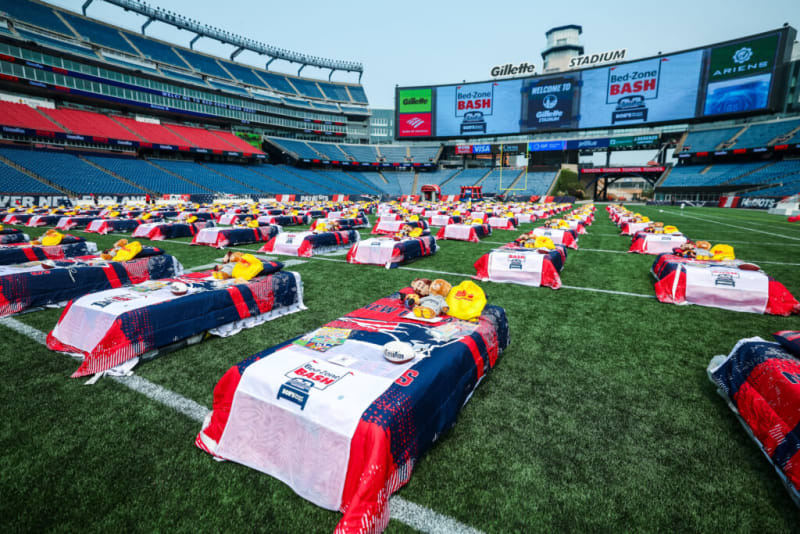 Gillette Stadium field set with multiple beds covered in New England Patriots-themed bedding and towels reading "Bed-Zone BASH," arranged for a promotional event.