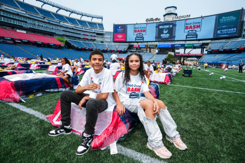 Two young people sitting on colorful bedding on a football field at Gillette Stadium during the Bed-Zone Bash event, wearing white Bed-Zone Bash t-shirts. The stadium seats and large digital screens with event branding are visible in the background.