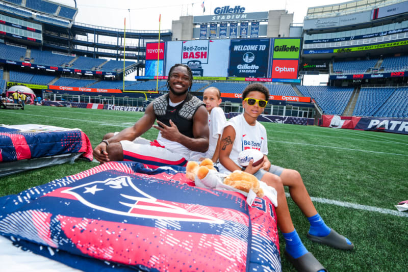 Three people sitting on a New England Patriots-themed field mattress inside Gillette Stadium with visible signage including "Bed-Zone BASH," "WELCOME TO Gillette STADIUM," "Toyota," "Optum," and "Gillette."
