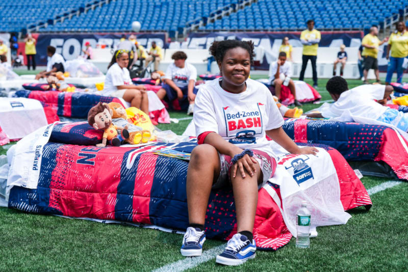 A child wearing a white "Bed-Zone BASH" t-shirt and sneakers sits on a red, blue, and white New England Patriots-themed bedspread with stuffed animals on a football field, surrounded by similar beds and children.