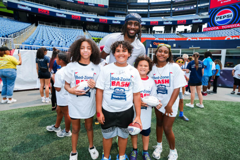 Group of children and an adult wearing white "Bed-Zone Bash" t-shirts holding footballs on a sports field inside a stadium with visible Pepsi and Toyota signage.