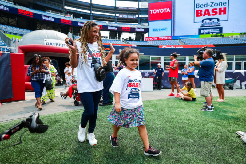 A woman and a child wearing white "Bed-Zone BASH" T-shirts walk on a stadium field with green artificial turf. The child wears a floral skirt and black sneakers. The woman carries a black crossbody bag and holds a phone. In the background, other people stand near a red inflatable tunnel and a large digital screen displays "Bed-Zone BASH," "TOYOTA," and "BUD LIGHT" logos, indicating a sponsored event at a sports stadium.