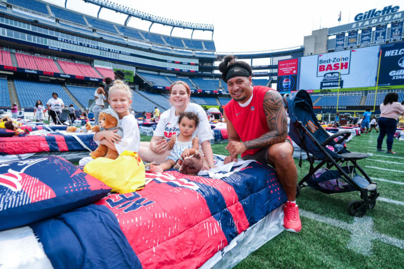 Family sitting on colorful bedding at Gillette Stadium during Bed-Zone Bash 2023 event with visible signage and stadium seating.
