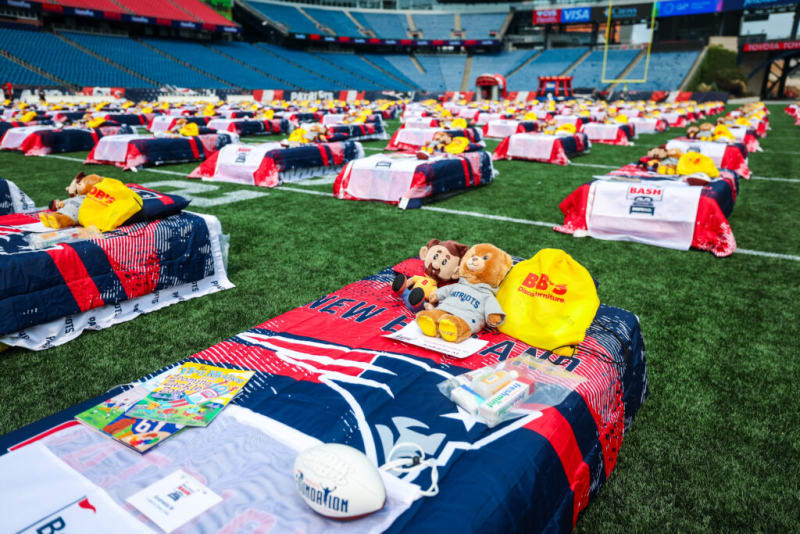 Beds with New England Patriots-themed bedding, stuffed animals, and yellow BB's Discount Furniture bags arranged on a football field for a charity event.