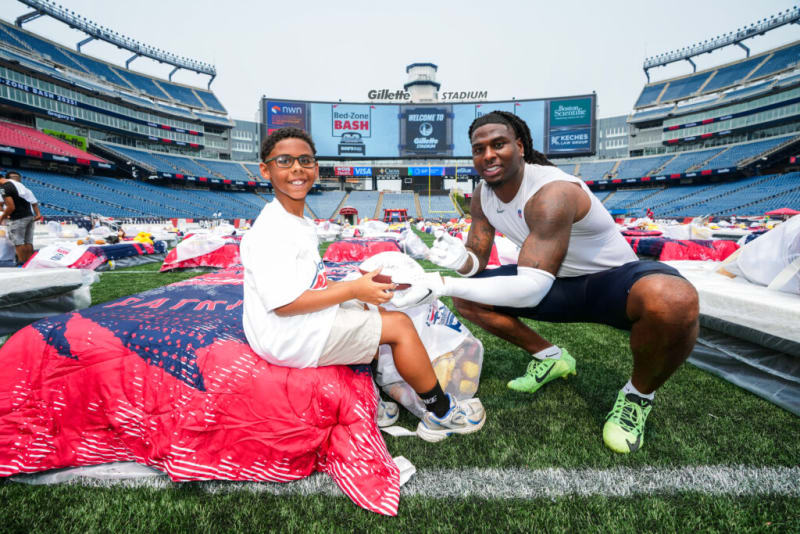 A young boy sitting on a red and blue sleeping bag on the field at Gillette Stadium, receiving a signed football from a football player in white and navy athletic gear with neon green cleats. The stadium scoreboard displays "WELCOME TO Gillette STADIUM" with sponsor logos.