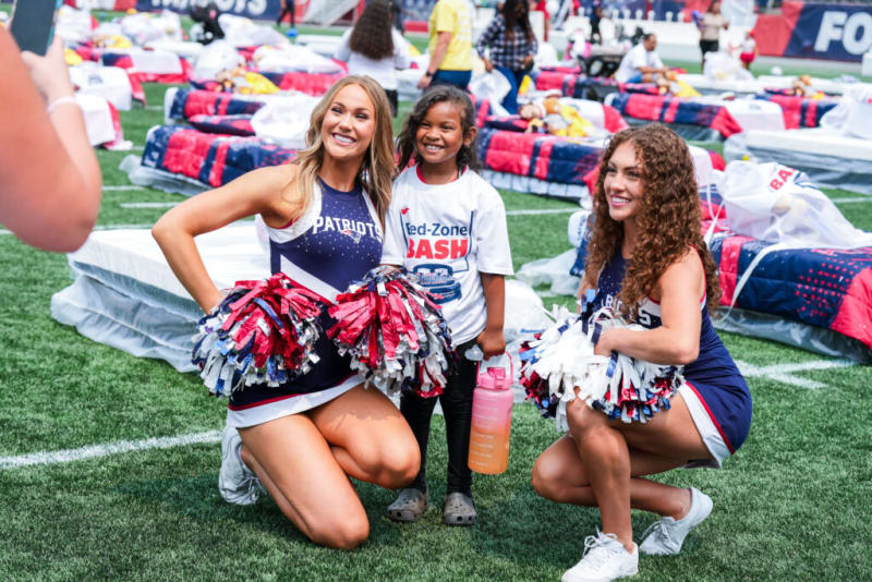 Two cheerleaders in navy blue Patriots uniforms with red, white, and blue pom-poms pose on a football field with a child wearing a white "Red-Zone Bash" t-shirt holding a pink water bottle.
