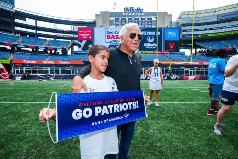 Two people at Gillette Stadium holding a blue banner that reads "Welcome to New England Go Patriots! Bank of America" on a football field with stadium seating in the background.