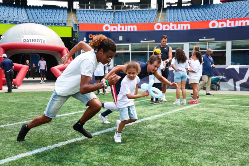 Young people running on a football field near a large inflatable Patriots helmet tunnel, with stadium seating and Optum branding visible.