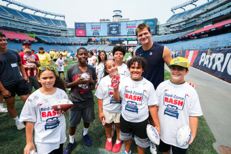 Group of children and an adult at Gillette Stadium wearing white "Bed-Zone BASH" t-shirts, holding footballs, participating in a football event.