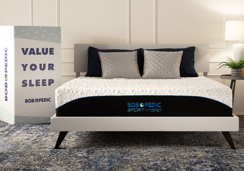 Modern bed with a Bob-O-Pedic Sport Hybrid mattress featuring a white quilted top and black sides with blue trim. Next to it is a large box with the text "VALUE YOUR SLEEP BOBOPEDIC" in bold navy letters. The bed has a light gray frame and is styled with four pillows in black and gray. The setting includes a blue and gray patterned rug, a side table with a lamp, and a plant, emphasizing comfort and quality sleep.