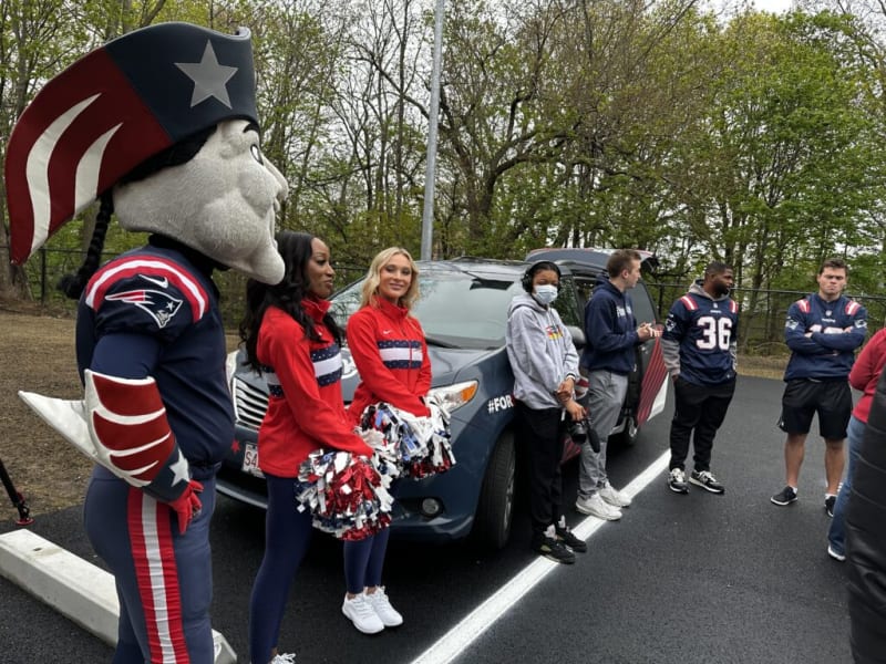 New England Patriots mascot and cheerleaders in red, white, and blue uniforms standing near a branded vehicle with fans wearing Patriots gear.