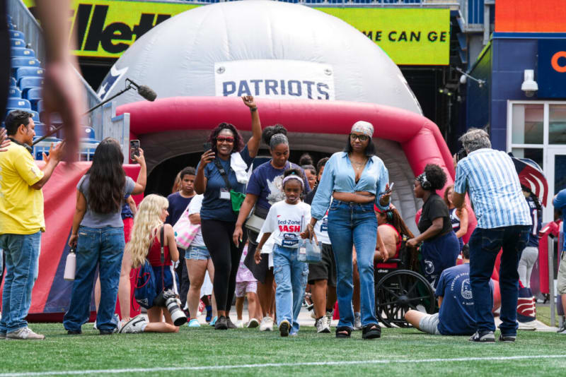 Group of diverse people walking on a football field near a large inflatable Patriots helmet tunnel, capturing a sports event atmosphere.