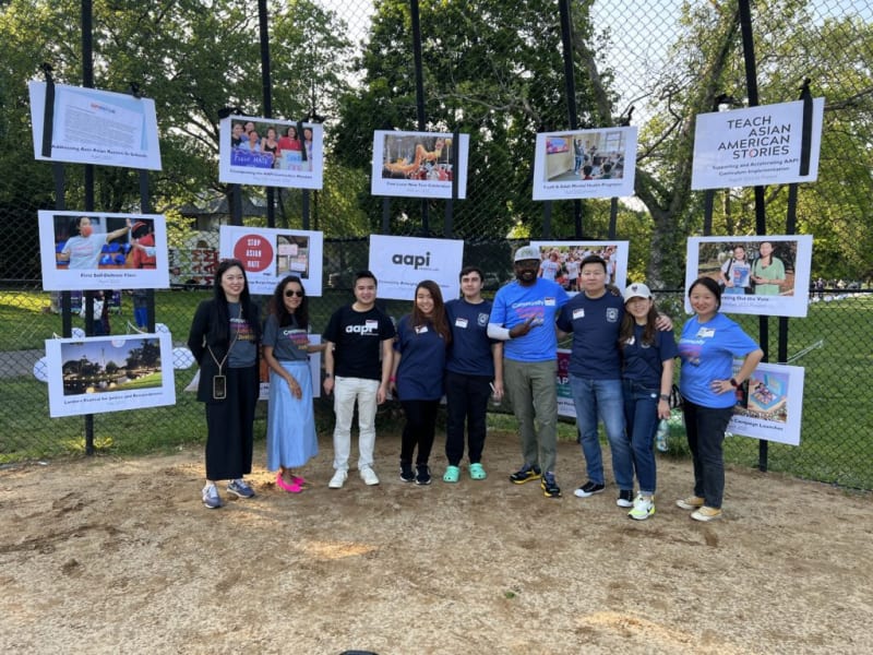 Group of people standing in front of a fence displaying posters on anti-Asian racism, mental health, voting, and community programs, promoting AAPI awareness and support. Posters include texts like "Addressing Anti-Asian Racism in Schools," "Stop Asian Hate," "Teach Asian American Stories," and "First Self-Defense Class."